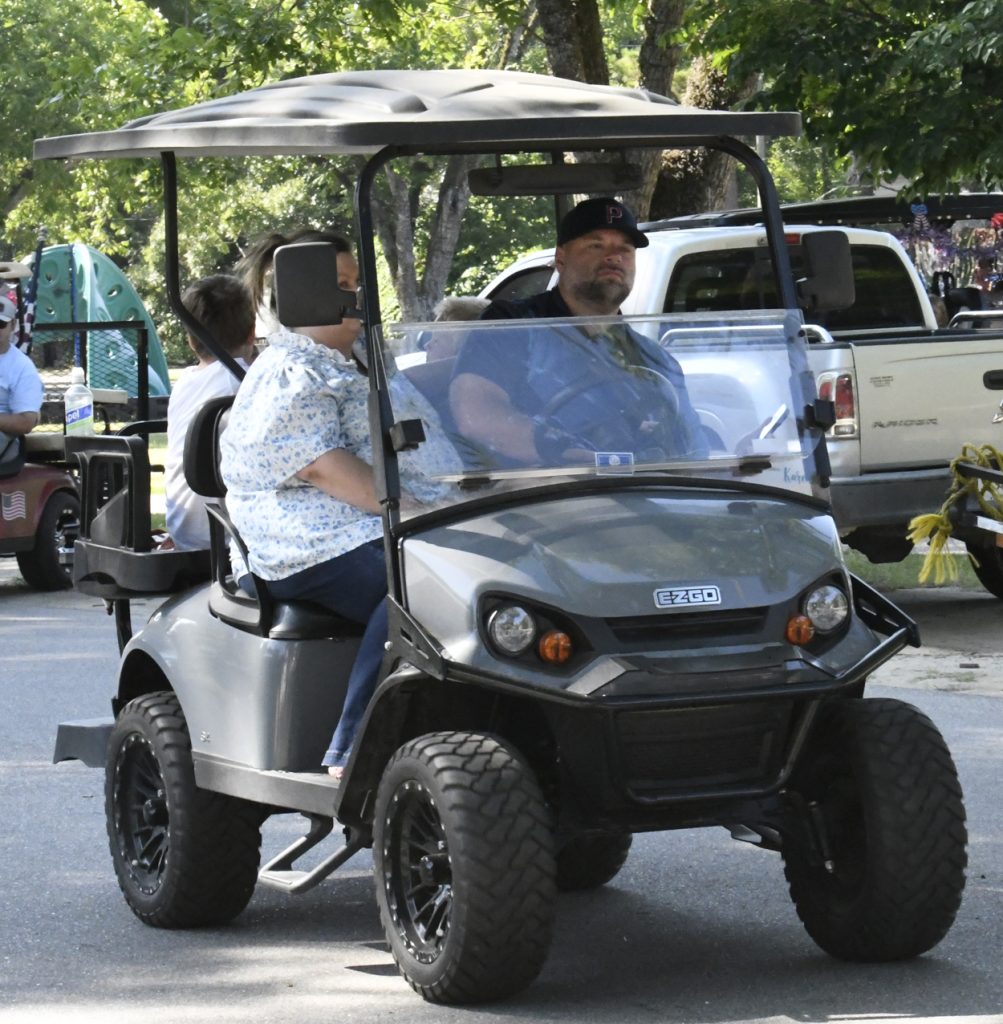 A man and woman ride in a black EZGO golf cart on a sunny day, with trees and parked vehicles in the background.