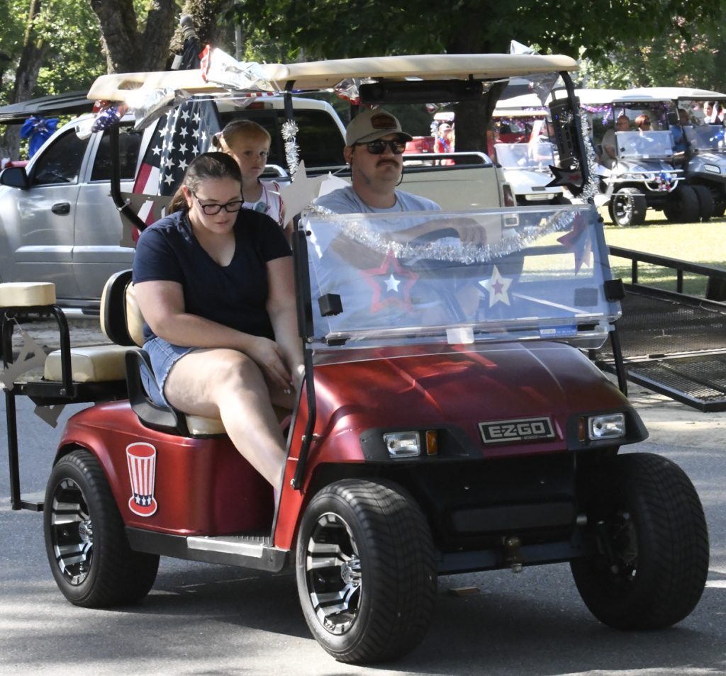 Three people ride a decorated red golf cart with patriotic stars and an American flag during a sunny outdoor event.