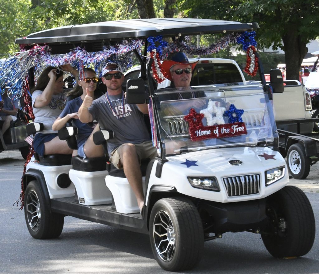 People riding a festively decorated golf cart with red, white, and blue tinsel and a "Home of the Free" sign, celebrating a patriotic event.