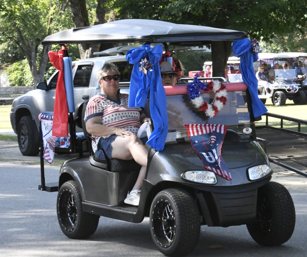 A decorated golf cart with patriotic USA flags and ribbons, driven by two people, participates in a parade with other decorated carts visible in the background.