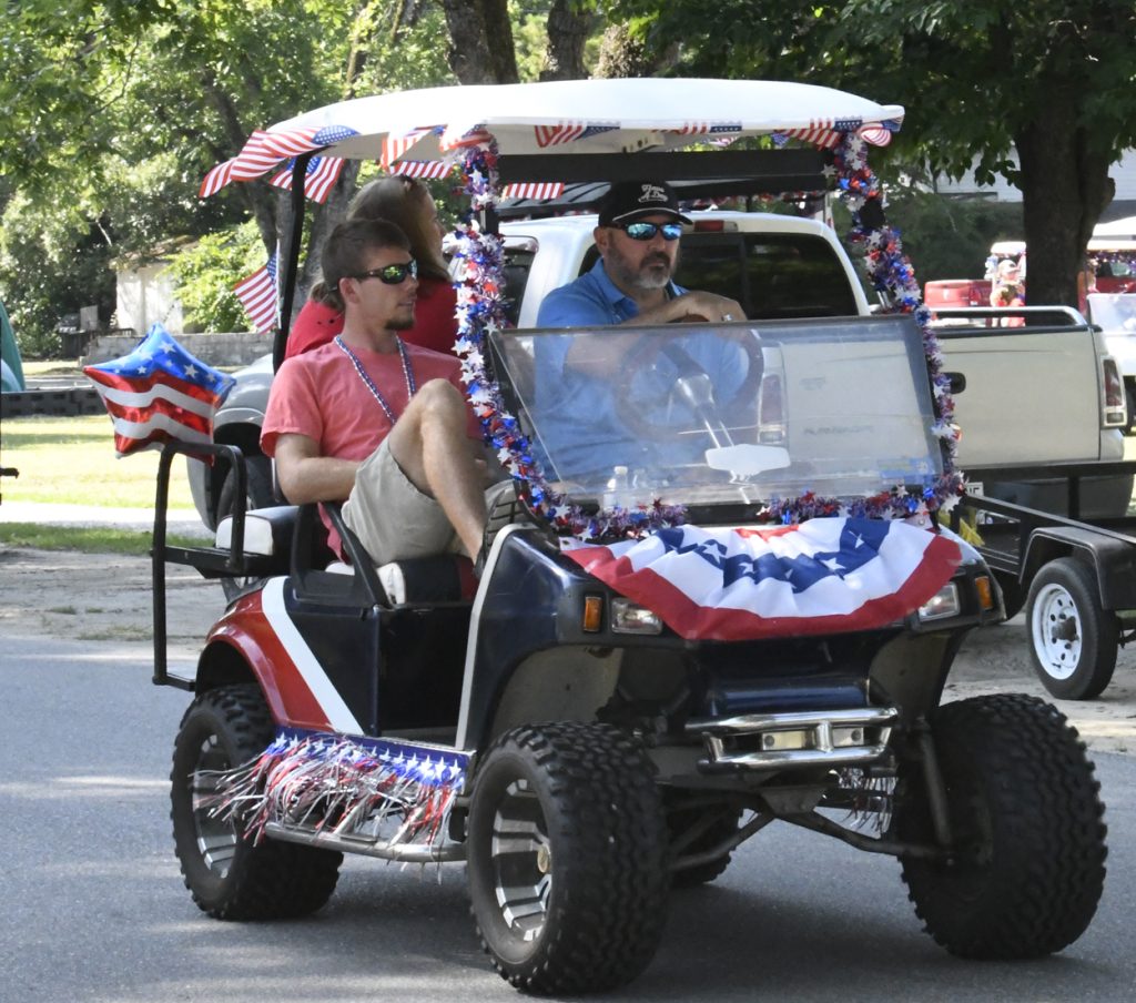 Two men ride a golf cart decorated with red, white, and blue patriotic decorations, including flags and garlands, during a parade or festive event.
