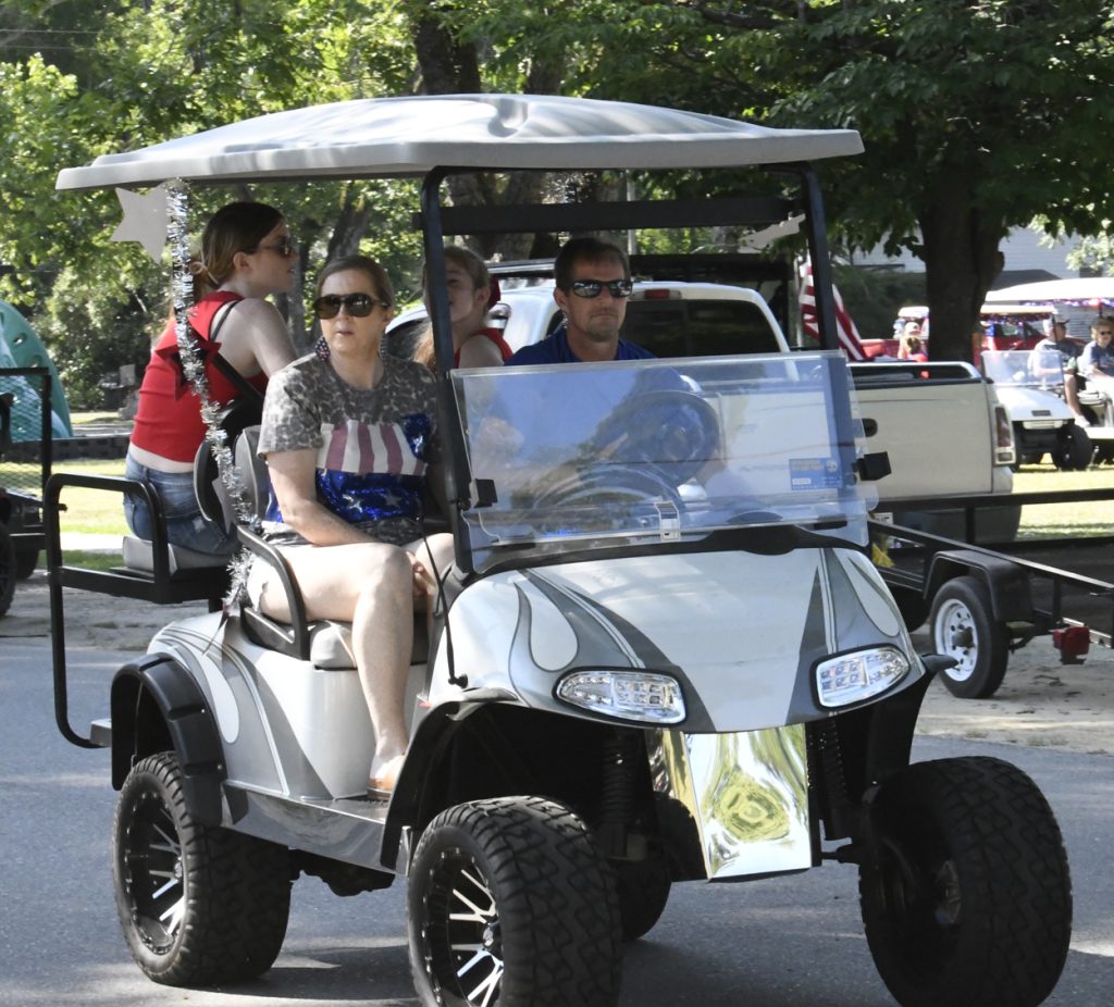Four people ride in a golf cart decorated for a festive event, driving down a street with trees and parked vehicles in the background.