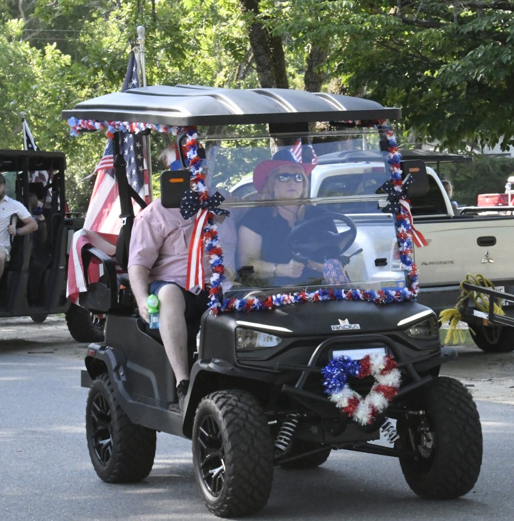 A golf cart decorated with American flags and red, white, and blue garlands drives down a street with two people participating in a parade.