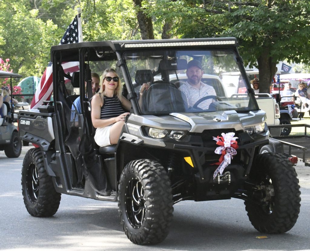 A man and woman ride in a decorated off-road vehicle with an American flag, participating in a festive parade on a sunny day.