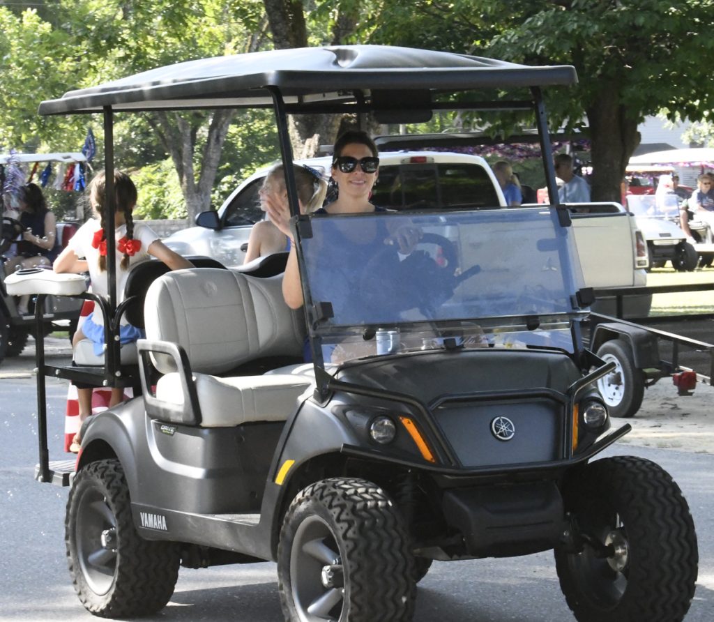 Woman driving a Yamaha golf cart with children riding in the back, outdoors in a sunny park with trees and several parked vehicles in the background.