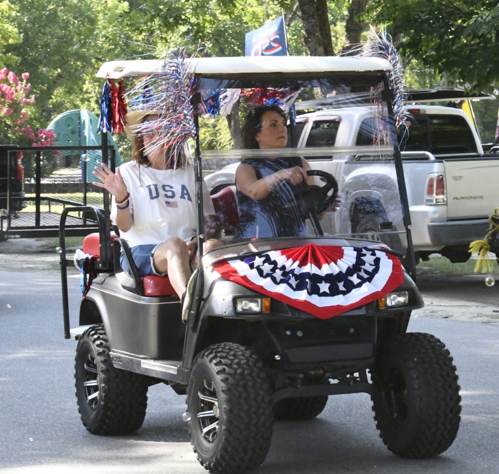Two women ride a golf cart decorated with red, white, and blue patriotic streamers and bunting, celebrating an American holiday, likely the Fourth of July.