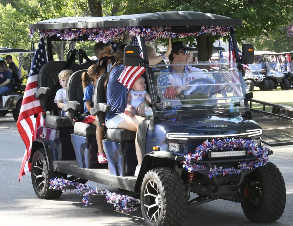 A decorated golf cart with American flags carries a family with children, participating in a parade under sunny, leafy trees.