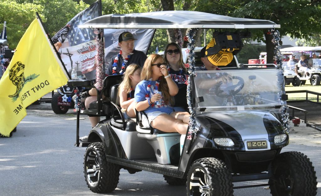 People riding a decorated golf cart in a parade, with a large yellow "Don't Tread On Me" flag and festive outfits, outdoors on a sunny day.