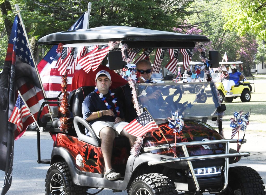 People riding a decorated golf cart with American flags and patriotic decorations, participating in what appears to be a parade on a sunny day.