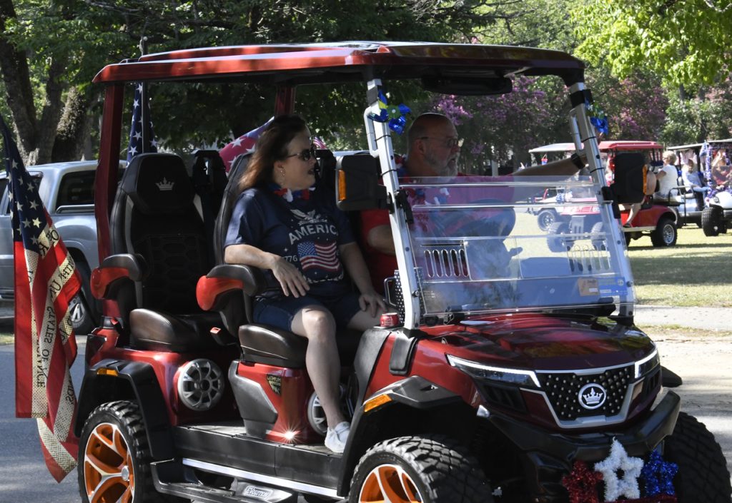 Two people ride in a decorated red golf cart with American flags, celebrating outdoors during a sunny day.