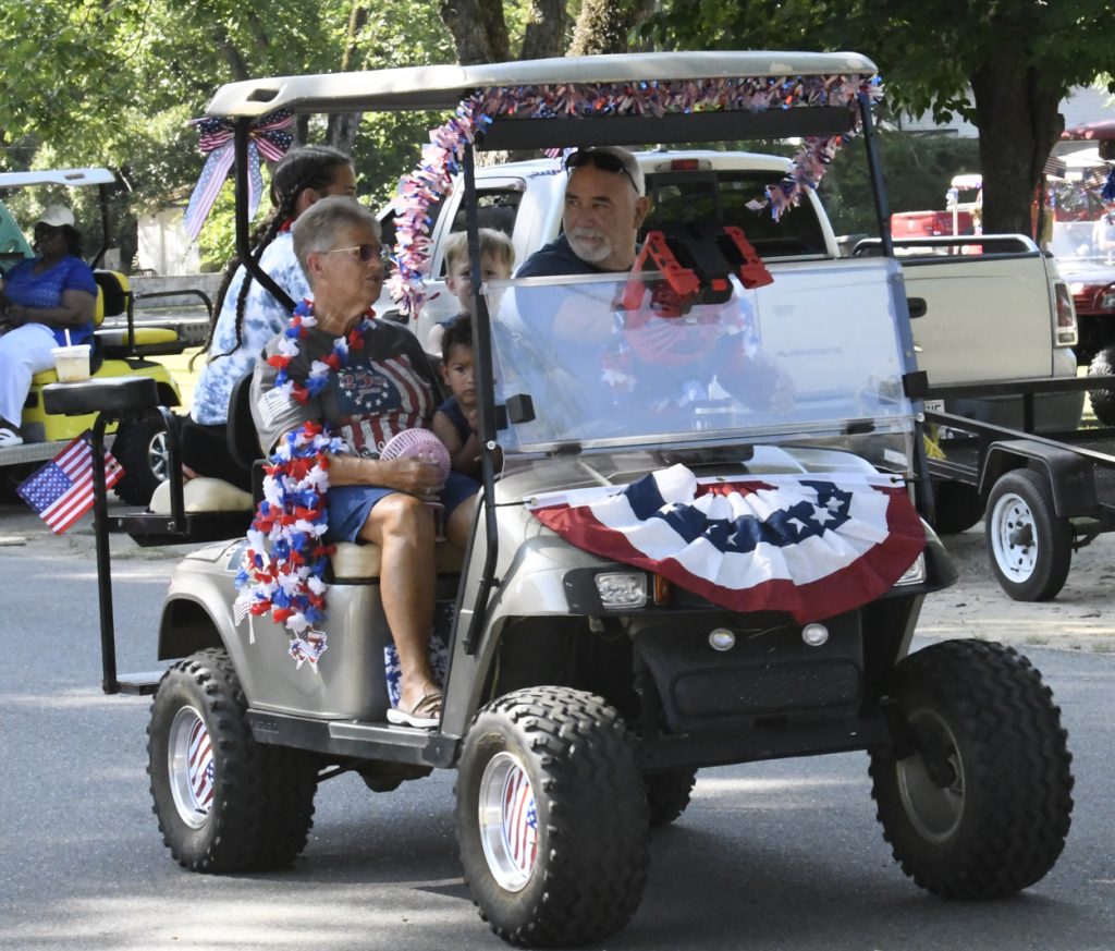 People riding a decorated golf cart with patriotic red, white, and blue decorations during a parade, American flags visible, summer outdoor setting.