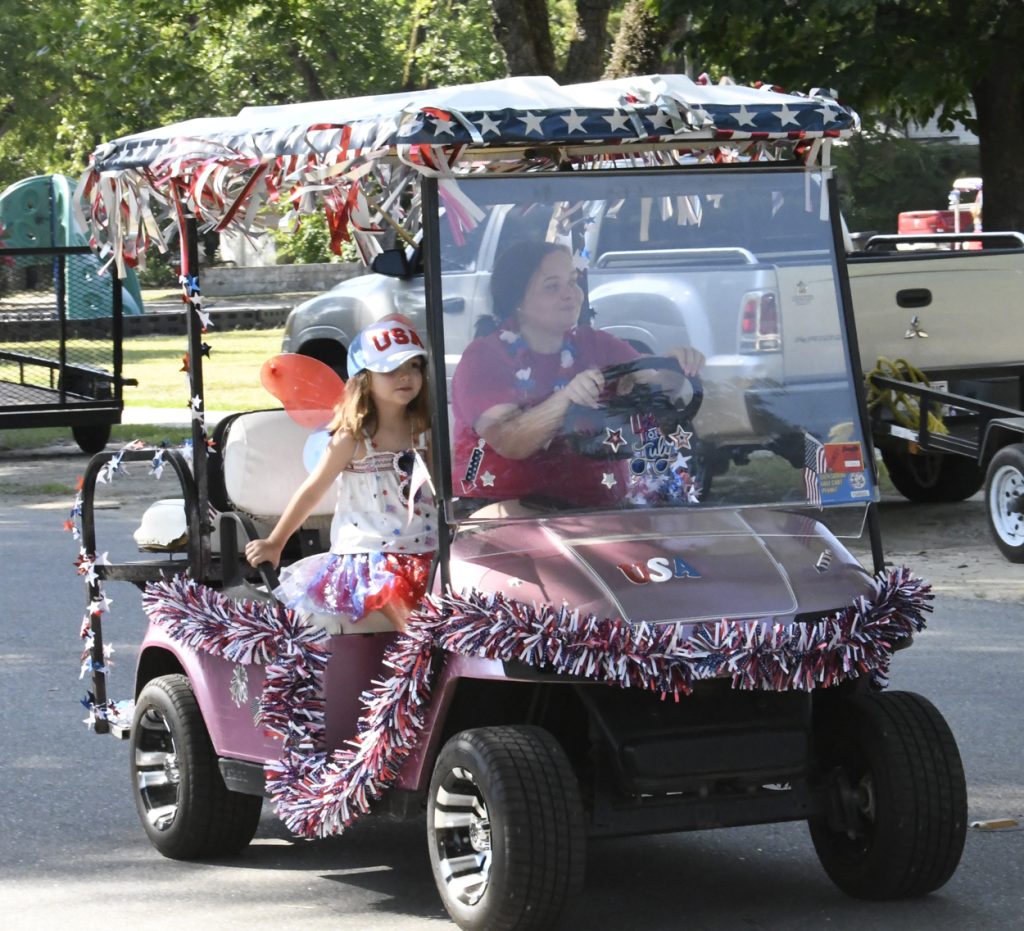 A woman and child ride a golf cart decorated with red, white, and blue streamers and USA symbols for a patriotic parade celebration.