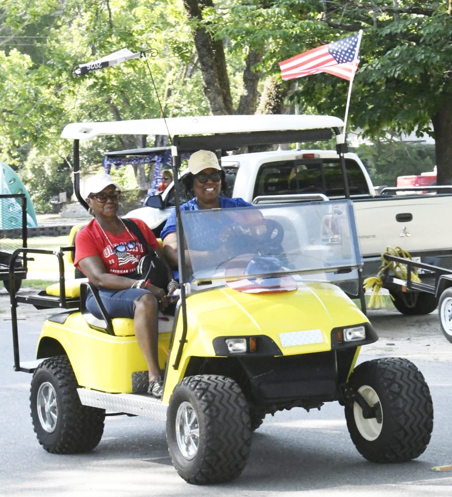 Two women ride a yellow golf cart decorated with an American flag, smiling on a sunny day with trees and parked vehicles in the background.