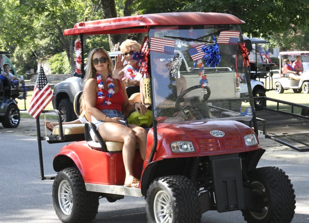 People riding a decorated red golf cart with American flags and patriotic garlands, celebrating outdoors on a sunny day.