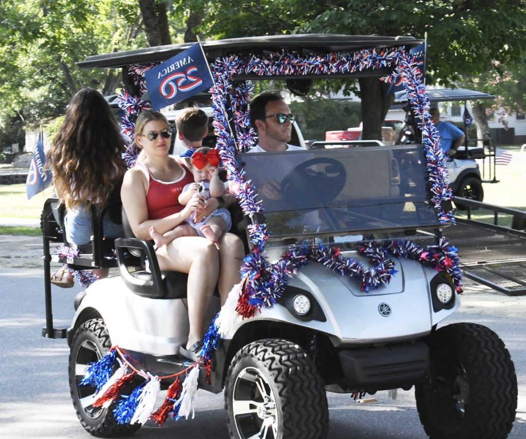 A family rides a decorated golf cart with American flags and red, white, and blue garlands during a parade on a sunny day.