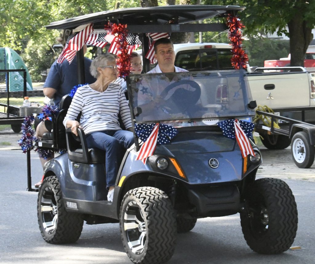 People riding a decorated golf cart with American flags and patriotic colors, participating in what appears to be a parade or festive event.