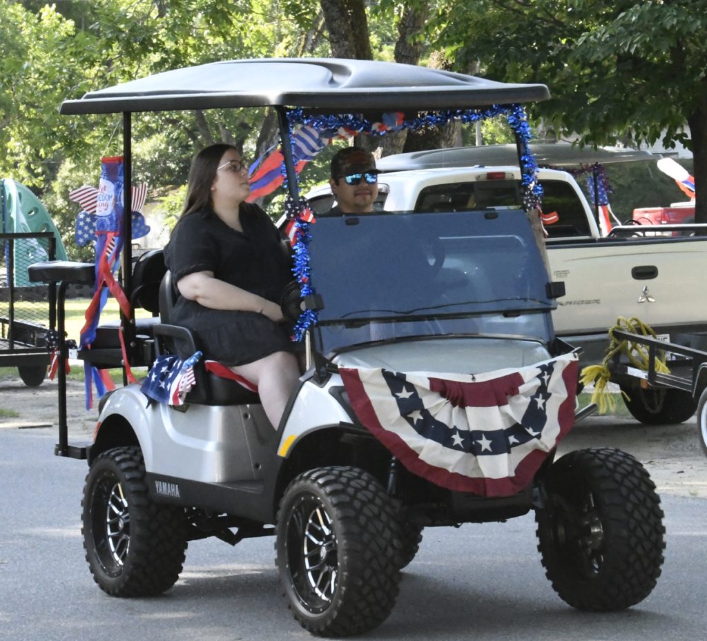 Two people ride a golf cart decorated with red, white, and blue garlands and banners for a patriotic celebration, with trees and vehicles in the background.