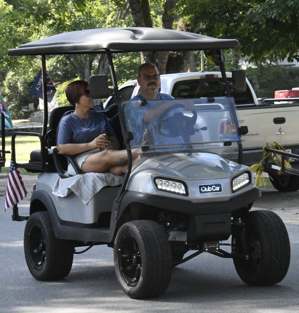 Two people ride in a gray golf cart on a sunny day, with an American flag attached to the cart and trees in the background.