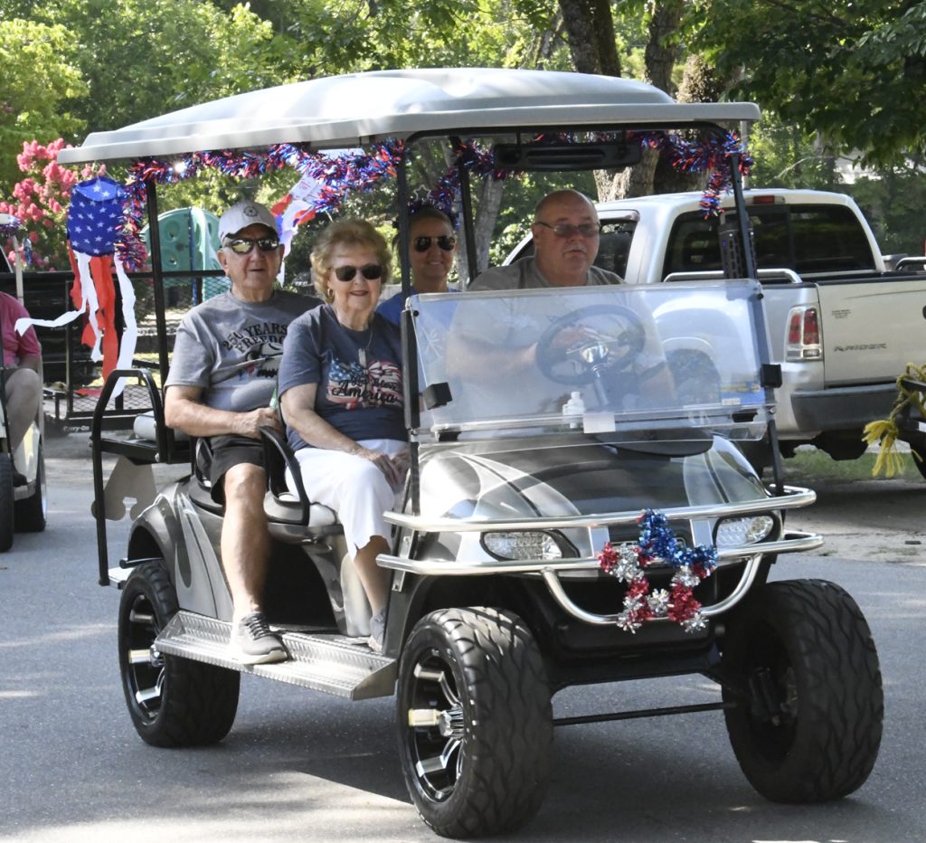 Four adults ride a decorated golf cart with patriotic red, white, and blue streamers, participating in an outdoor event under sunny, leafy trees.