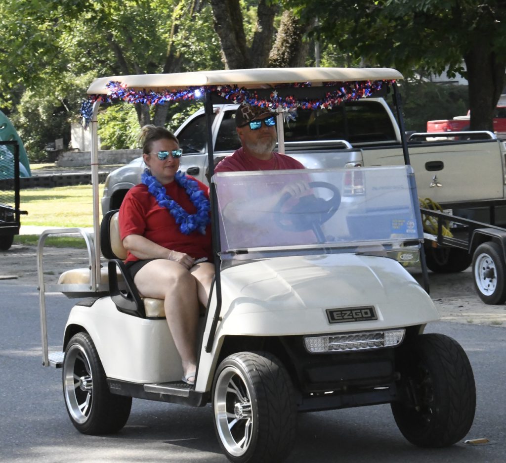 Two people wearing sunglasses and red shirts ride a decorated golf cart on a sunny day, surrounded by parked vehicles and trees.