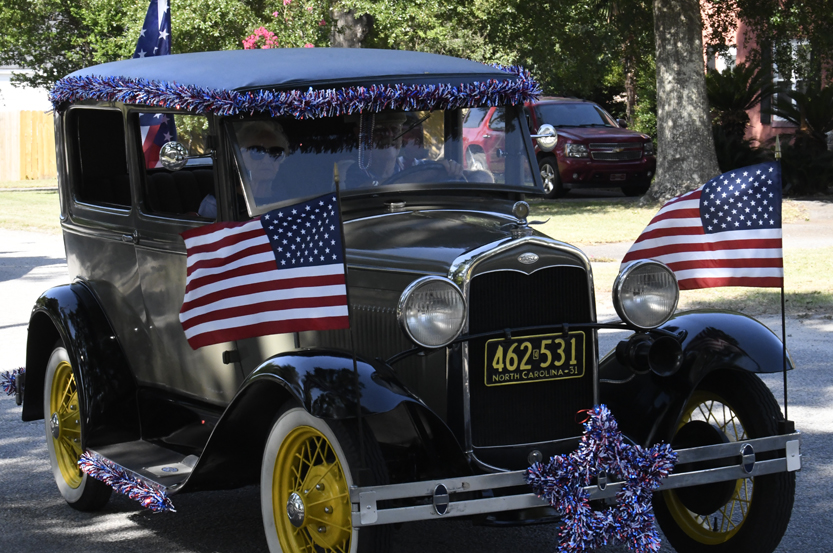 Vintage car decorated with American flags and patriotic garlands, participating in a parade on a sunny day.