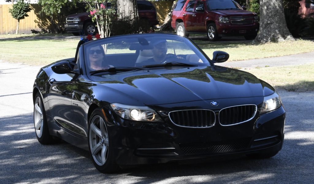 A black BMW convertible with two people inside driving on a residential street, trees and parked cars visible in the background.