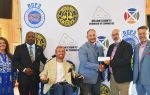 Six people posing for a photo, one man in a wheelchair, two men shaking hands and holding a check, with a Dillon County Chamber of Commerce backdrop.