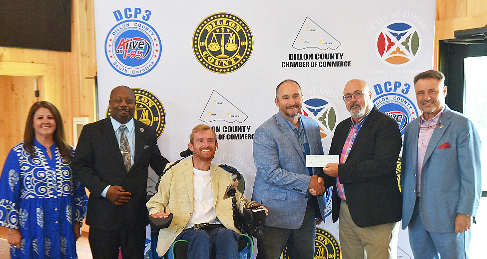 Six people posing for a photo, one man in a wheelchair, two men shaking hands and holding a check, with a Dillon County Chamber of Commerce backdrop.