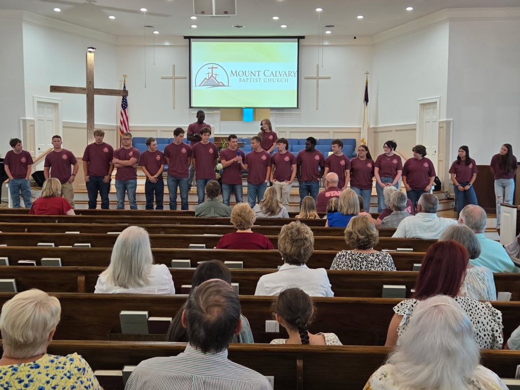 A group of young people in matching maroon shirts stand at the front of a church, facing an audience seated in pews.
