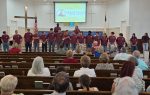 A group of young people in matching maroon shirts stand at the front of a church, facing an audience seated in pews.
