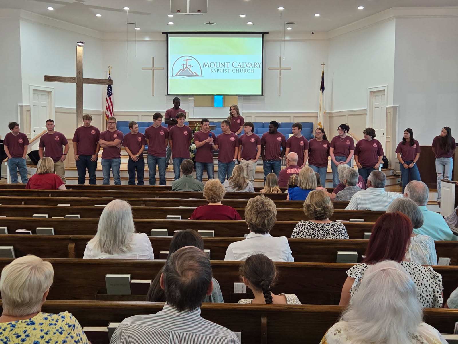 A group of young people in matching maroon shirts stand at the front of a church, facing an audience seated in pews.