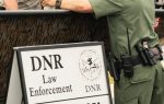 A wildlife officer talks to a man in a boat beside a sign that reads "DNR Law Enforcement Courtesy Boat Inspections" with an arrow pointing right.