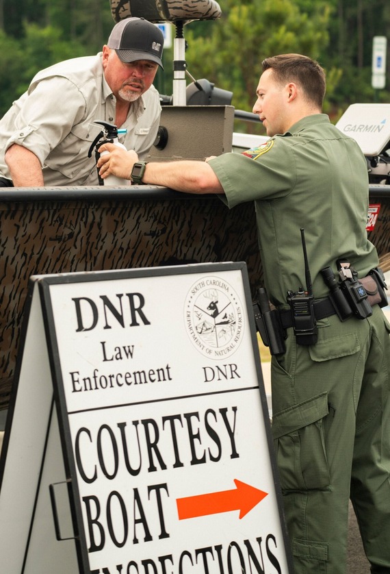 A wildlife officer talks to a man in a boat beside a sign that reads "DNR Law Enforcement Courtesy Boat Inspections" with an arrow pointing right.