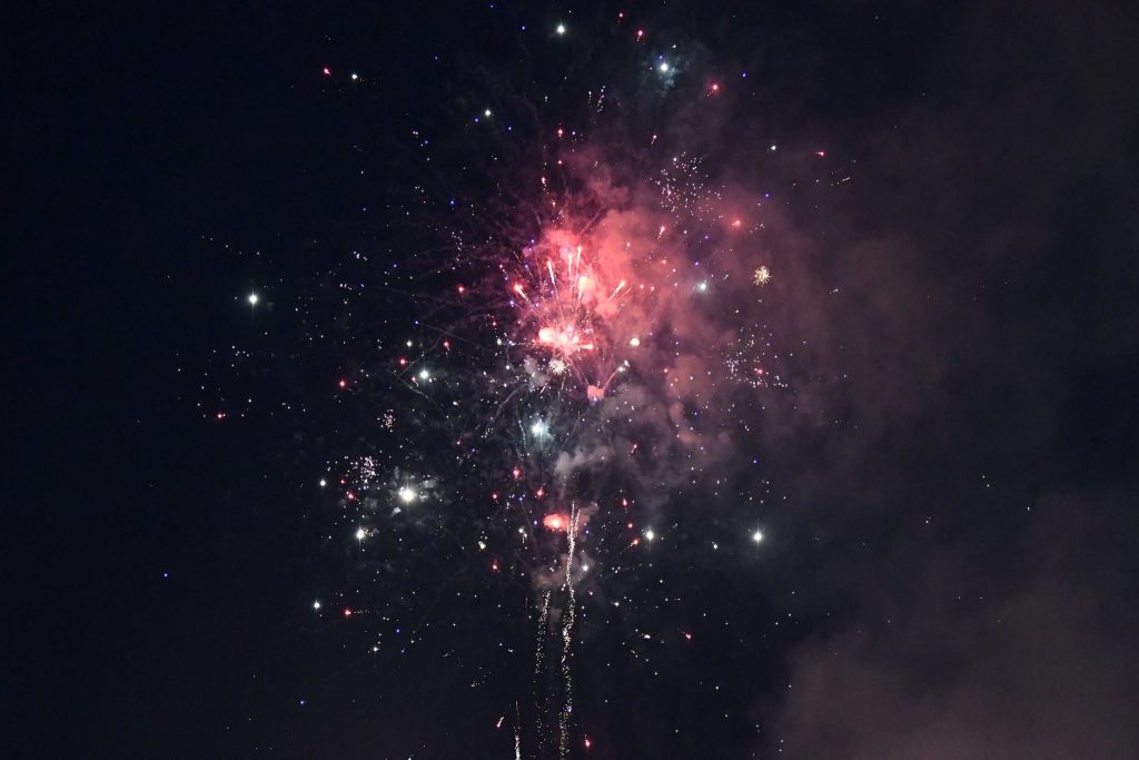 Colorful fireworks explode in the night sky, creating bursts of red, white, and blue light, with smoke and sparkles surrounding the display.
