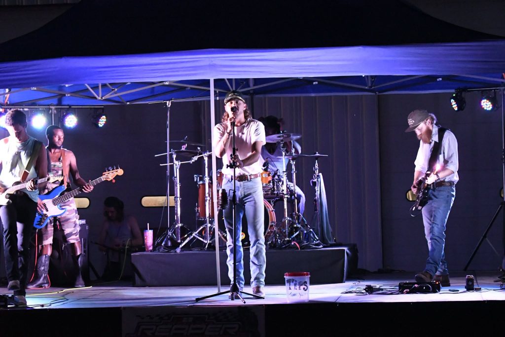 A band performs live on an outdoor stage under blue canopy lights, with musicians playing guitar, bass, drums, and a vocalist at the center microphone.
