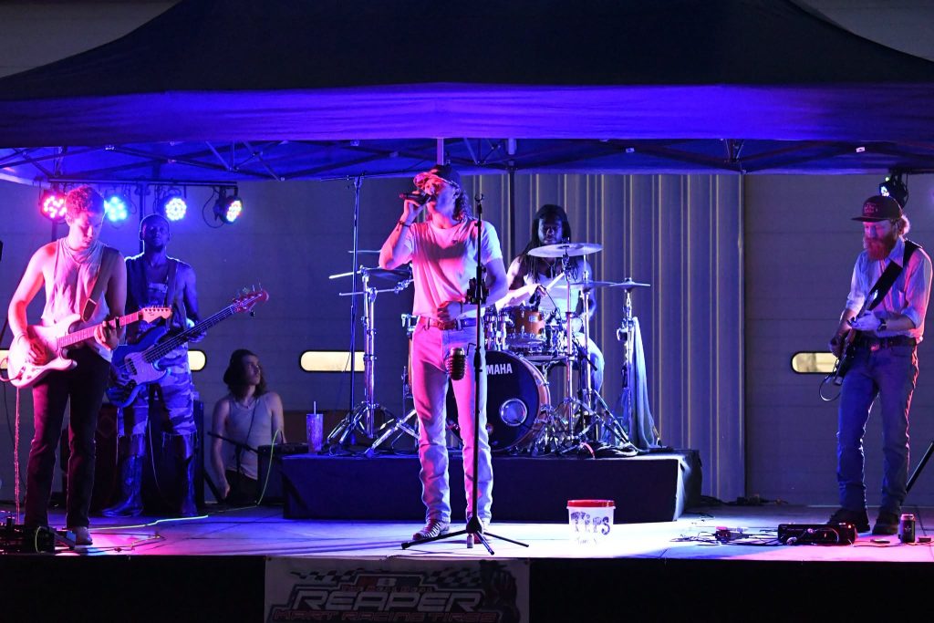 A band performs live on an outdoor stage at night, with colorful lights and a visible tip jar in front of the singer.
