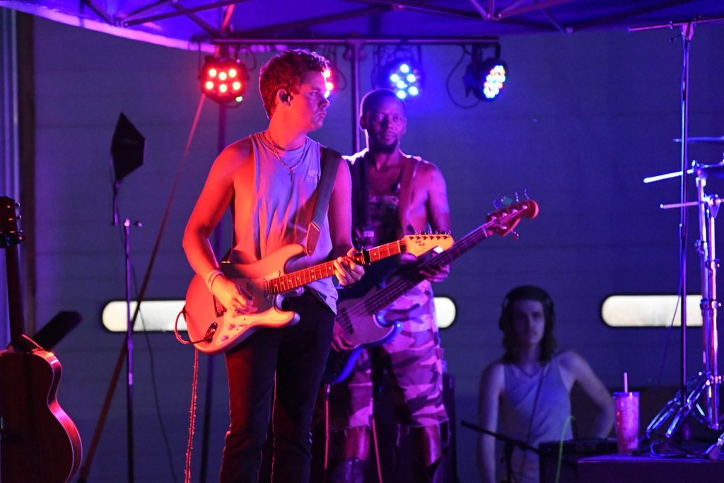Two musicians perform on stage under colorful lights, one playing electric guitar and another playing bass, with a third person in the background wearing headphones.