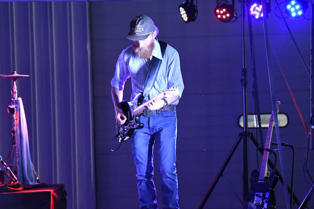 Man in casual clothes and cap playing electric guitar on stage under colorful lights, with another guitar and drum set visible in the background.