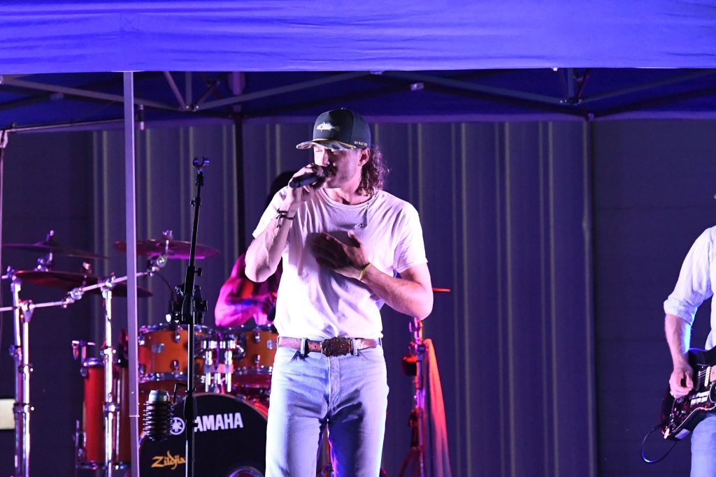 A man in a white shirt and cap sings into a microphone on stage, with a drummer and guitarist behind him.