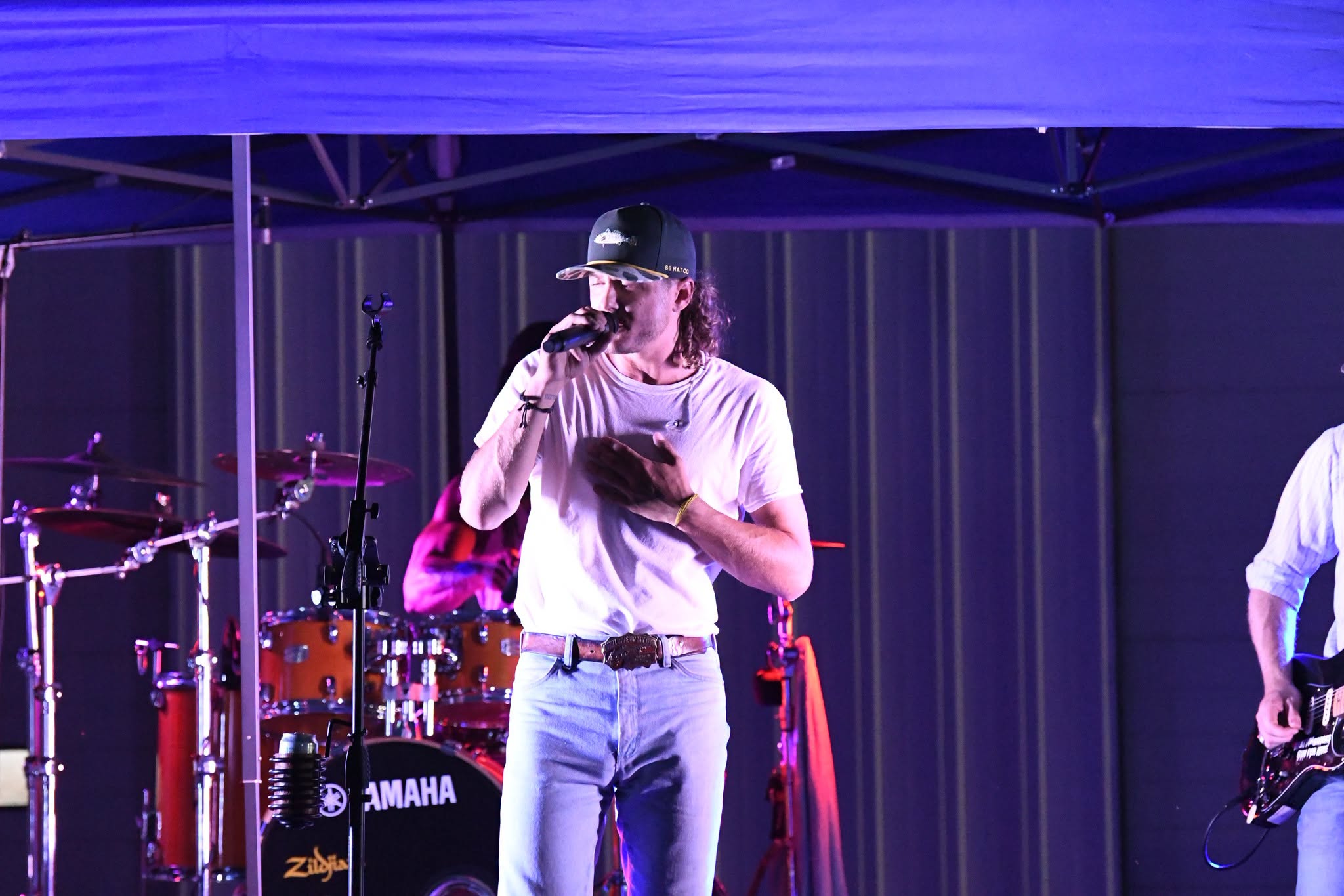 A man in a white shirt and cap sings into a microphone on stage, with a drummer and guitarist behind him.