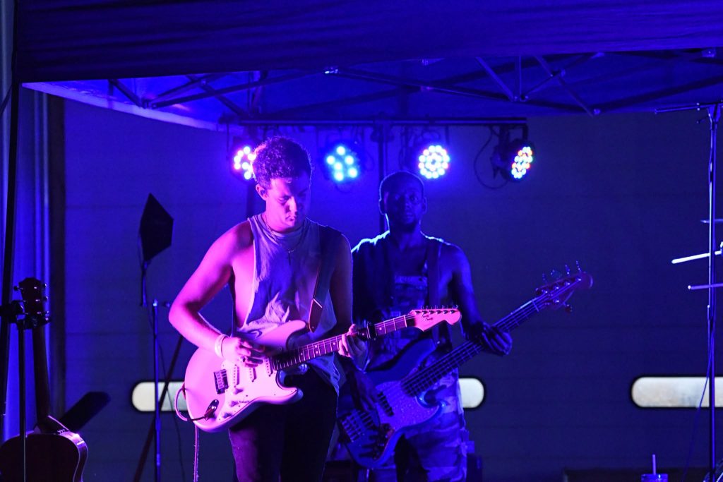 Two musicians perform on stage under purple and blue lights, one playing electric guitar and the other playing bass guitar, with stage equipment visible in the background.