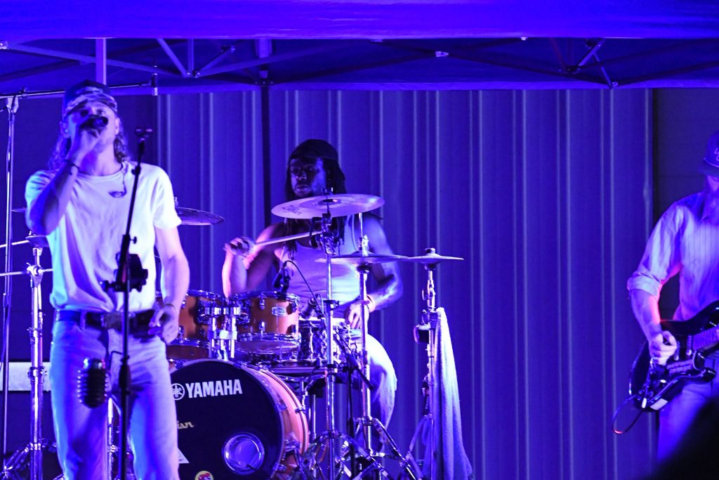 A band performs on stage under purple lighting, featuring a singer, drummer, and guitarist, with a Yamaha drum set visible.