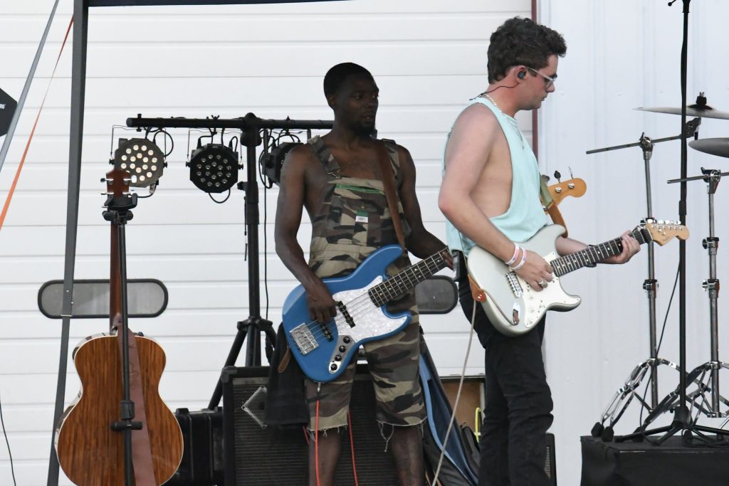 Two musicians perform on stage, one playing a blue electric bass guitar and the other playing a white electric guitar, with musical equipment and lights behind them.