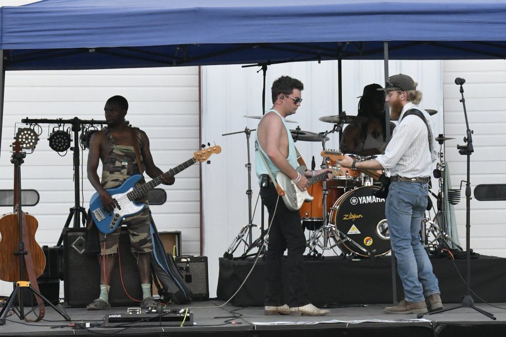 Four musicians perform on an outdoor stage under a blue canopy, playing electric guitar, bass, and drums, with instruments and equipment visible.
