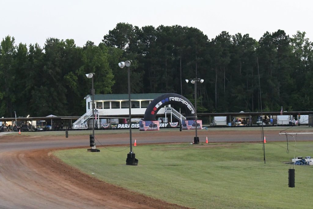 Dirt racetrack with lighting poles, a white control building, "PARADISE WAY" sign, inflatable archway, and trees in the background.