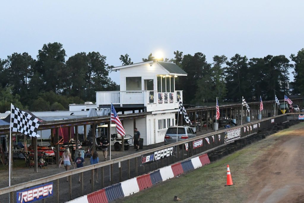 A small race track pit area with an elevated building, checkered flags, American flags, parked vehicles, people walking, and trees in the background at dusk.