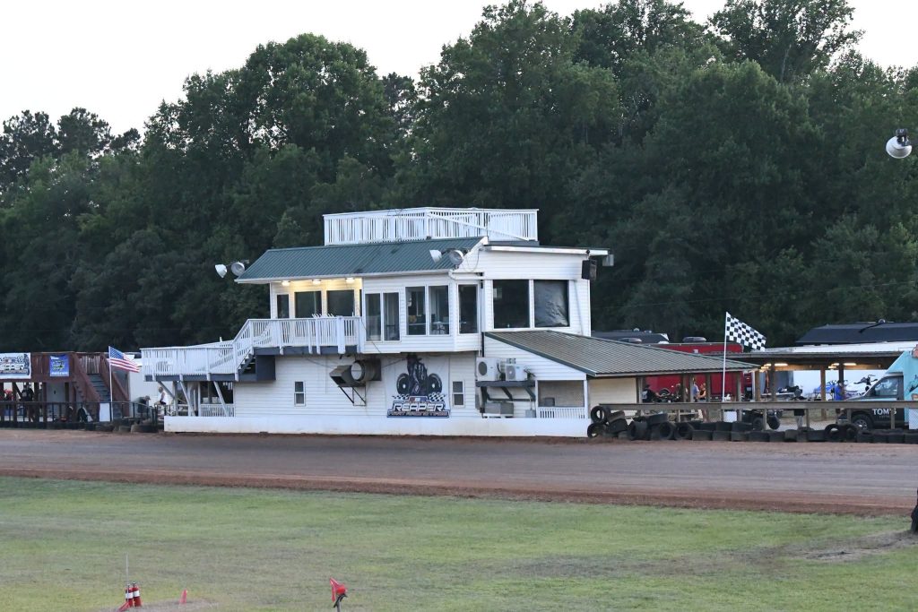A white announcer’s booth with large windows overlooks a dirt racetrack. Behind it are trees and a checkered flag. Racing vehicles are parked nearby.