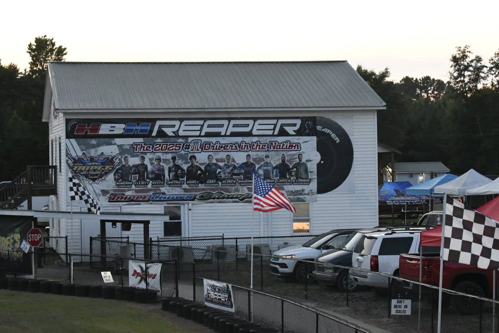 A raceway building with banners showing drivers, an American flag, parked cars, tents, and checkered flags in the foreground.