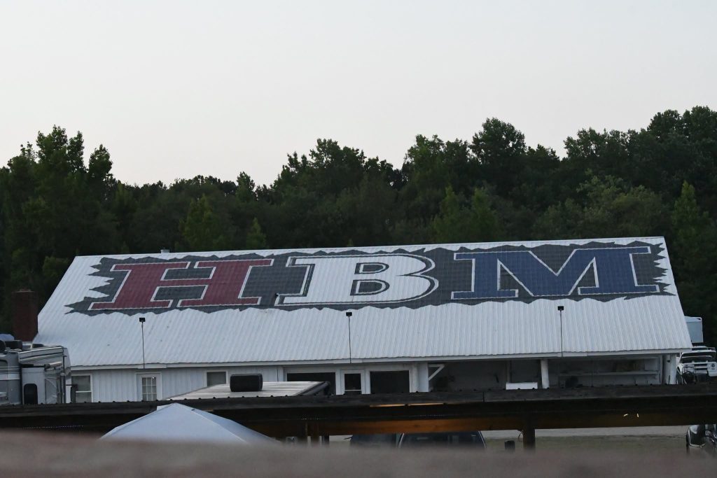 Large white roof with big, stylized letters "HBM" painted in red, white, and blue, surrounded by trees in the background.