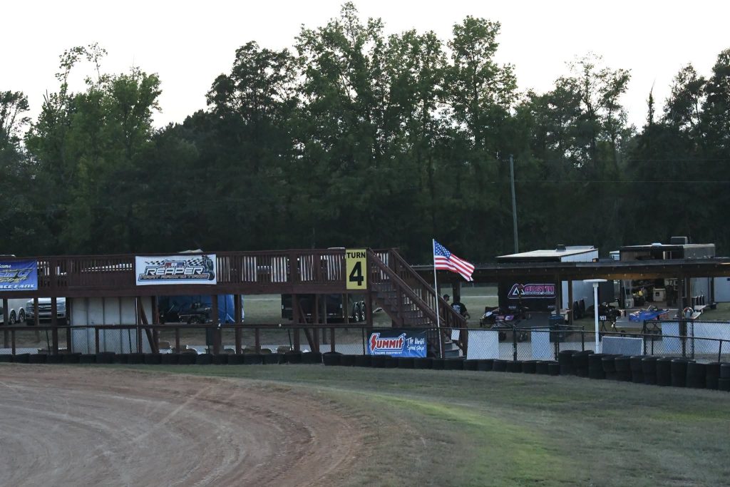 Dirt racetrack curve near grandstand with Turn 4 sign, American flag, trailers, and trees in the background.